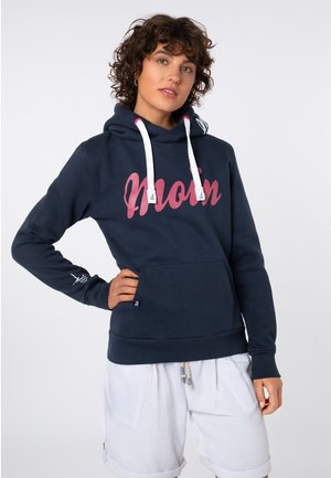 Young person with curly hair wearing navy blue hoodie with pink "Mom" text and white shorts, posing with one hand on hip against plain background.