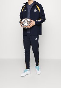 Persoon draagt een marineblauw Real Madrid trainingspak en witte voetbalschoenen, houdt een zwart-witte voetbal met oranje details vast.