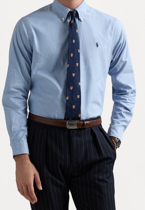 Man draagt een lichtblauw overhemd, een marineblauwe stropdas met een geel schildpatroon, een bruine riem en zwarte pantalon met krijtstreep, met één hand in zijn broekzak.