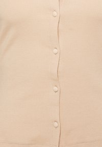 Beige ribbad kofta med en rad av sex tygklädda knappar fram och en rundad nedre kant. Slät textur med subtil stretch.