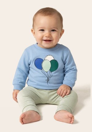 Smejoč dojenček sedi bosa, oblečen v modro kapucarico z motivom balona in svetlo zelene hlače na enobarvnem ozadju.