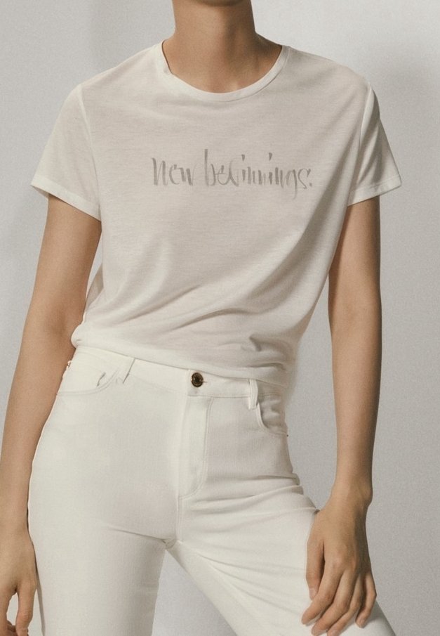 Personne portant un t-shirt blanc avec le texte « Nouveaux départs » et un pantalon blanc taille haute, debout avec une main posée sur la cuisse.