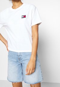Vit bomulls-T-shirt med korta ärmar, prydd med en liten Tommy Jeans-logotyp i röd, svart och vit på vänster bröst. Matchad med blå denimshorts.