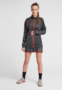 Tenue de sport comprenant une veste zippée grise avec des accents orange et des manches à motifs en chevrons, associée à des shorts assortis et des sneakers.