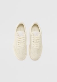 Sneaker beige in suede con una texture liscia, punta arrotondata, lacci piatti e logo sulla linguetta. Design minimalista con suole in gomma.