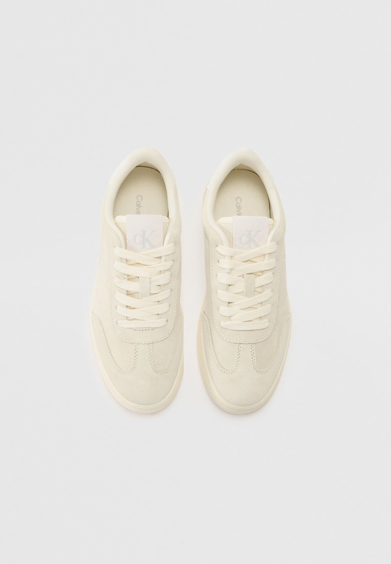 Beżowe zamszowe sneakersy o gładkiej teksturze, zaokrąglonym czubku, płaskich sznurowadłach i logo na języku. Minimalistyczny design z gumowymi podeszwami.