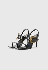 Black leather high-heeled sandals featuring open toe, ankle strap, and gold hardware accents. Smooth texture and minimalist design.