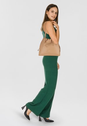 Beige Bucket Bag aus glattem Leder mit Kordelzugverschluss und geprägtem Logo. Modell trägt einen grünen Overall und schwarze High Heels.