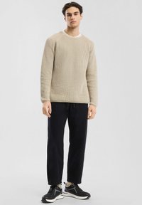 Jongeman staat in een bewerkte beige trui, zwarte broek en zwarte sneakers met witte zolen op een effen lichte achtergrond.