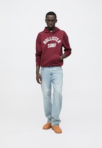 Hanorac bordo cu textul „HOLLISTER SURF”, blugi albaștri și pantofi slip-on maro deschis, pe un fundal neutru. Design și croială simple.