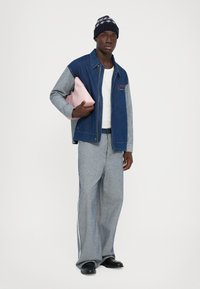 Homme portant une veste en denim bleu et gris, un pantalon assorti, une chemise blanche, des chaussures noires, un bonnet en tricot, tenant une pochette rose sous le bras, se tenant devant un fond blanc.