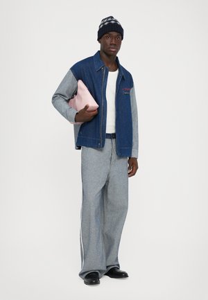 Homme portant une veste en denim bleu et gris, un pantalon assorti, une chemise blanche, des chaussures noires, un bonnet en tricot, tenant une pochette rose sous le bras, se tenant devant un fond blanc.