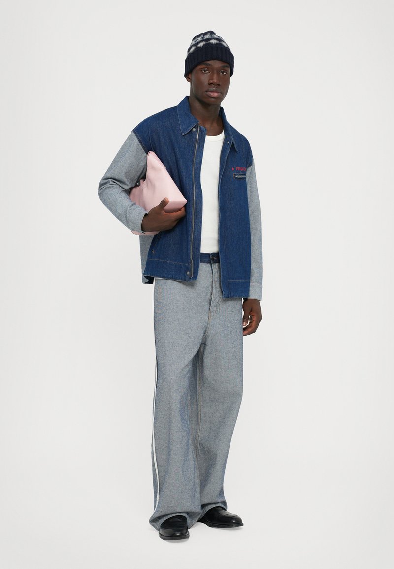 Homme portant une veste en denim bleu et gris, un pantalon assorti, une chemise blanche, des chaussures noires, un bonnet en tricot, tenant une pochette rose sous le bras, se tenant devant un fond blanc.