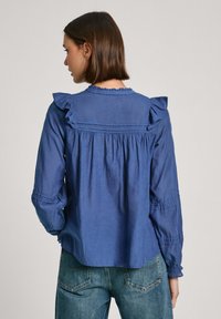 Blusa leggera blu con dettagli arruffati sulle spalle, retro arricciato, maniche lunghe e vestibilità comoda. Abbinata a jeans di denim.