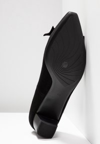 Chaussure à talon en daim noir inclinée contre un mur, montrant la semelle avec une adhérence texturée et un talon carré. Taille indiquée 42½ sur la semelle.