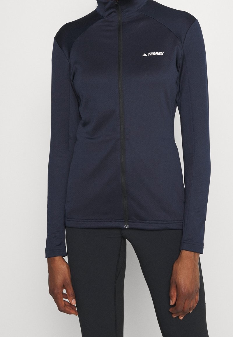 Veste de sport Terrex bleu marine zippée intégralement portée avec un legging noir moulant sur une personne debout sur un fond uni.