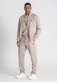 Beige trainingspak met logo-patroon, inclusief een zipjack en joggingsbroek met trekkoord. Witte sneakers maken de outfit compleet. Rode tekstaccenten.