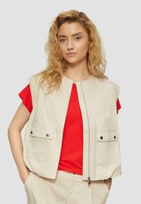 Beige ärmellose Weste mit Reißverschluss vorne, zwei Fronttaschen und Druckknopfverschlüssen. Getragen über einem roten T-Shirt mit glatter Textur.