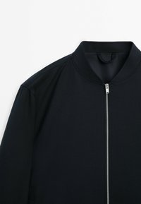 Zwarte jas met opstaande kraag en rits aan de voorkant. Gemaakt van gladde stof, met gestructureerde schouders en een strak lijnenontwerp.