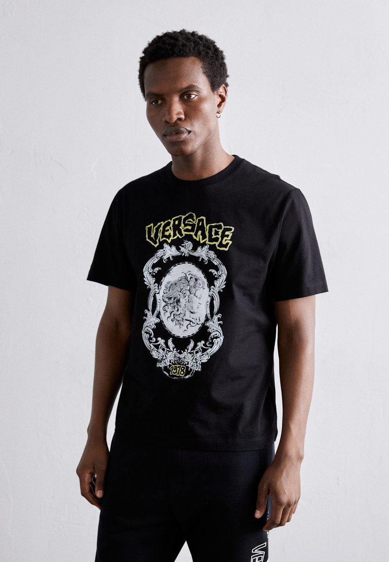 Must natuke mustast puuvillast, millel on suur graafika näost, mis on kaunistatud keeruka äärisega ning millel on kollane tekst "Versace". Lühikesed varrukad, ümar kaelus.