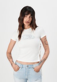 White short-sleeve t-shirt with a fitted design, featuring a glossy logo on the front and lace-up accents on the sides.