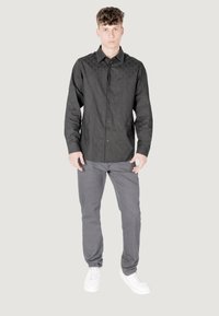 Camisetta nera a maniche lunghe con un motivo geometrico testurizzato, abbinata a pantaloni grigi con fantasia e sneakers bianche, in piedi davanti a uno sfondo neutro.