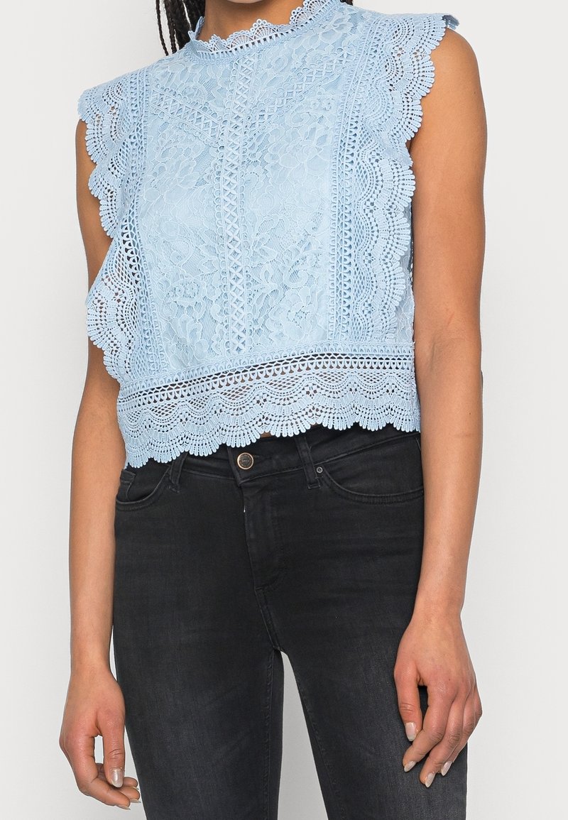 Ljusblå spetsblus med vågiga ärmslut och nederkant, intrikata blommönster och hög krage, kombinerad med mörka jeans.