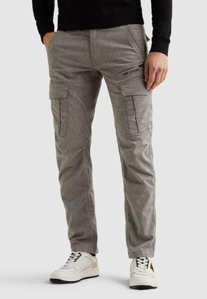 Muž v sivých nákladných nohaviciach s bočnými vreckami a bielych teniskách, stojaci so založenými rukami v vreckách na jednoduchom pozadí.