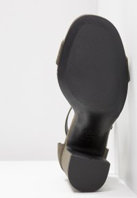 Vue de la semelle d'une chaussure à talon haut en daim taupe avec semelle extérieure en caoutchouc noir texturé et talon bloc, inclinée sur un fond blanc.
