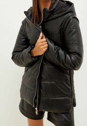 Vrouw draagt een zwarte gewatteerde leren jas met capuchon en bijpassende zwarte leren korte broek, is de jas gedeeltelijk aan het dichtritsen.