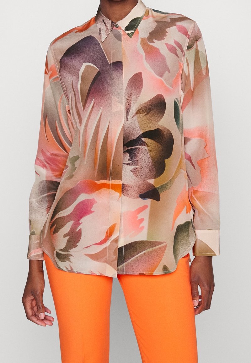 Chemise à manches longues avec col, confectionnée dans un tissu doux, arborant un motif floral aux tons roses, oranges et verts. Associée à un pantalon orange pour un look complet.