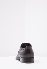 Chaussure habillée en cuir noir avec une texture lisse, un bout arrondi, et des détails discrets de brogue le long des coutures ; dotée d'un petit talon empilé.