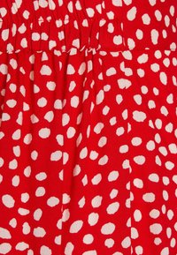 Červená tkanina s nabíranou texturou, se vzorem bílých puntíků. Zdůrazňuje hladký, lehký materiál.