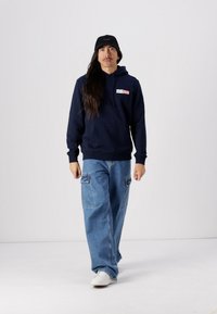 Mørkeblå hettegenser med logo-patch, parret med lyseblå baggy jeans med cargo-lommer. Hvite sneakers fullfører antrekket.