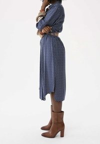 Femme portant une robe bleue à motifs et des bottes à talons mi-mollet marron, se tenant de profil avec les bras croisés et une main touchant son menton.