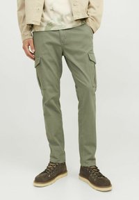 Pantalones cargo verdes con bolsillos laterales; fabricados en material de algodón; corte ajustado; textura lisa; combinados con botas marrones de cordones.