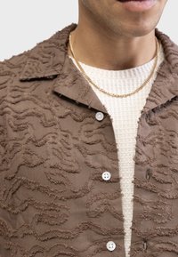 Brown shirt with textured, wavy pattern and white buttons; layered over a light cream knit top. Gold chain necklace around the neck.