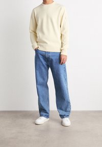 Světlý žlutý svetr s kulatým výstřihem, žebrovanými manžetami a kapsou na zip. Kombinován se světlými modrými volnými džíny a bílými teniskami.