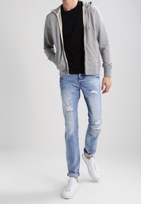 Szara bluza z kapturem zapinana na zamek na czarnej koszulce, zestawiona z przetartymi jasnoniebieskimi dżinsami i białymi trampkami. Luźna stylizacja, minimalne detale.