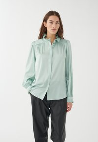 Hellgrüne Bluse mit Rüschensaum am Kragen und an den Manschetten, langen Puffärmeln und Knopfverschluss. Getragen mit schwarzer, locker sitzender Hose.