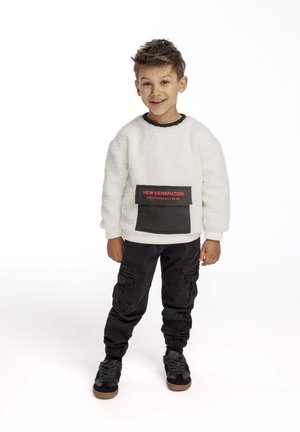 Vit, fluffig sweatshirt med en svart framlapp och röd text, i kombination med svarta cargobyxor och svarta sneakers med bruna sulor.