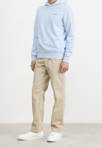 Felpa di cotone azzurra con tasca frontale e logo, abbinata a chino beige e sneakers bianche, in piedi su un pavimento neutro.