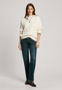 Pinti baltas megztinis su tekstūruotu deimanto raštu, užtrauktuku ant apykaklės ir raukinių rankogaliais, derinamas su tamsiai mėlynais tiesiais džinsais ir rudos spalvos batais.