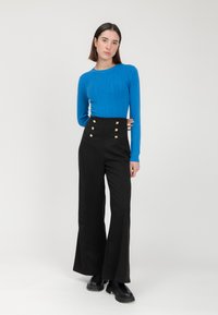 Pantalones negros de talle alto con botones dorados y piernas acampanadas, combinados con un suéter azul ajustado y de canalé. Tela suave, diseño moderno.