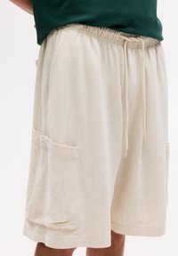 Shorts en coton beige avec une taille élastique, un cordon de serrage et deux poches latérales surdimensionnées. Texture douce avec une coupe décontractée.