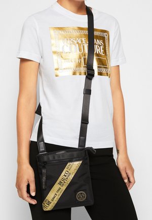 Osoba nosící bílé tričko se zlatým logem Versace Jeans Couture a černou kabelku přes rameno se zlatým diagonálním proužkem loga.