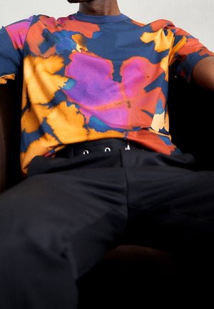 Mann sitzend, trägt ein buntes Batikshirt mit orange-, lila-, rot- und blau gemusterten Farben und schwarze Hosen mit Ösen am Bund.