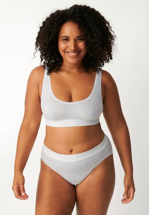 Donna sorridente con capelli ricci che indossa un reggiseno grigio chiaro e mutandine abbinate, in piedi davanti a uno sfondo bianco uniforme.