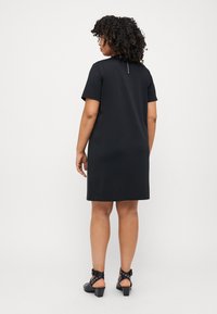 Black short-sleeve dress with a straight cut, smooth texture, and a back zip closure. Paired with black heeled sandals featuring cutouts.