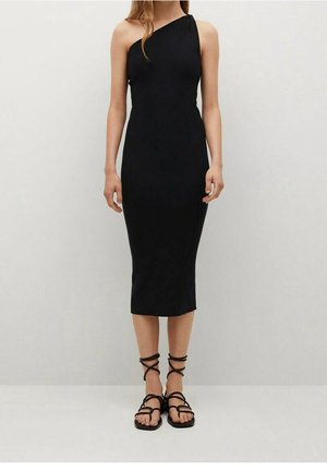 Kobieta w czarnej, midi sukience na jedno ramię bez rękawów oraz czarnych, płaskich sandałach na paski, stojąca na tle jasnego, jednolitego tła.